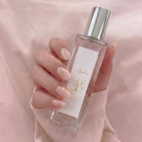 Maison de TracéのフレグランスAzalea。淡いピンクの背景に映える、上品なラベルが貼られた透明な香水ボトルを持つ手元。
